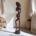 Handgesneden houten beeld van een gestileerde zwangere Afrikaanse vrouw met langgerekte vormen, een licht gebogen houding en een kamvormige hoofdtooi met ingesneden patronen.
