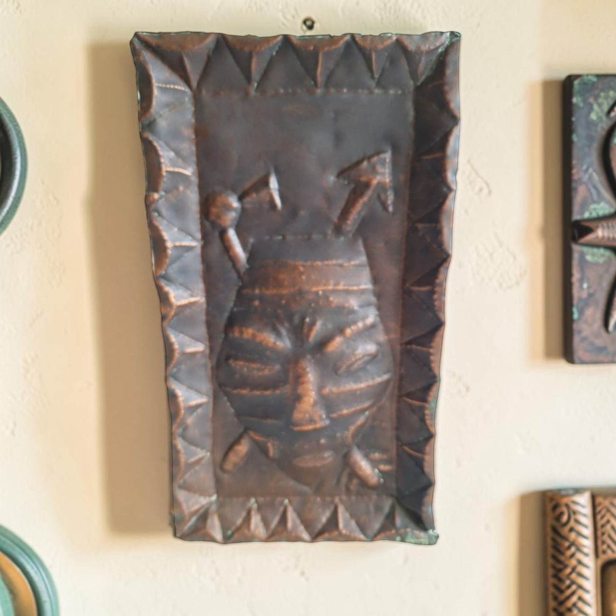 Koperkleurige Afrikaanse wandplaat met een gestileerd masker en geometrische patronen.
