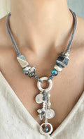 Halsketting met grijs suède koord, blauwe kristallen en facetgeslepen hematietkralen, afgewerkt met een cascade van zilveren ringen, muntjes en een blauwe steenhanger.
