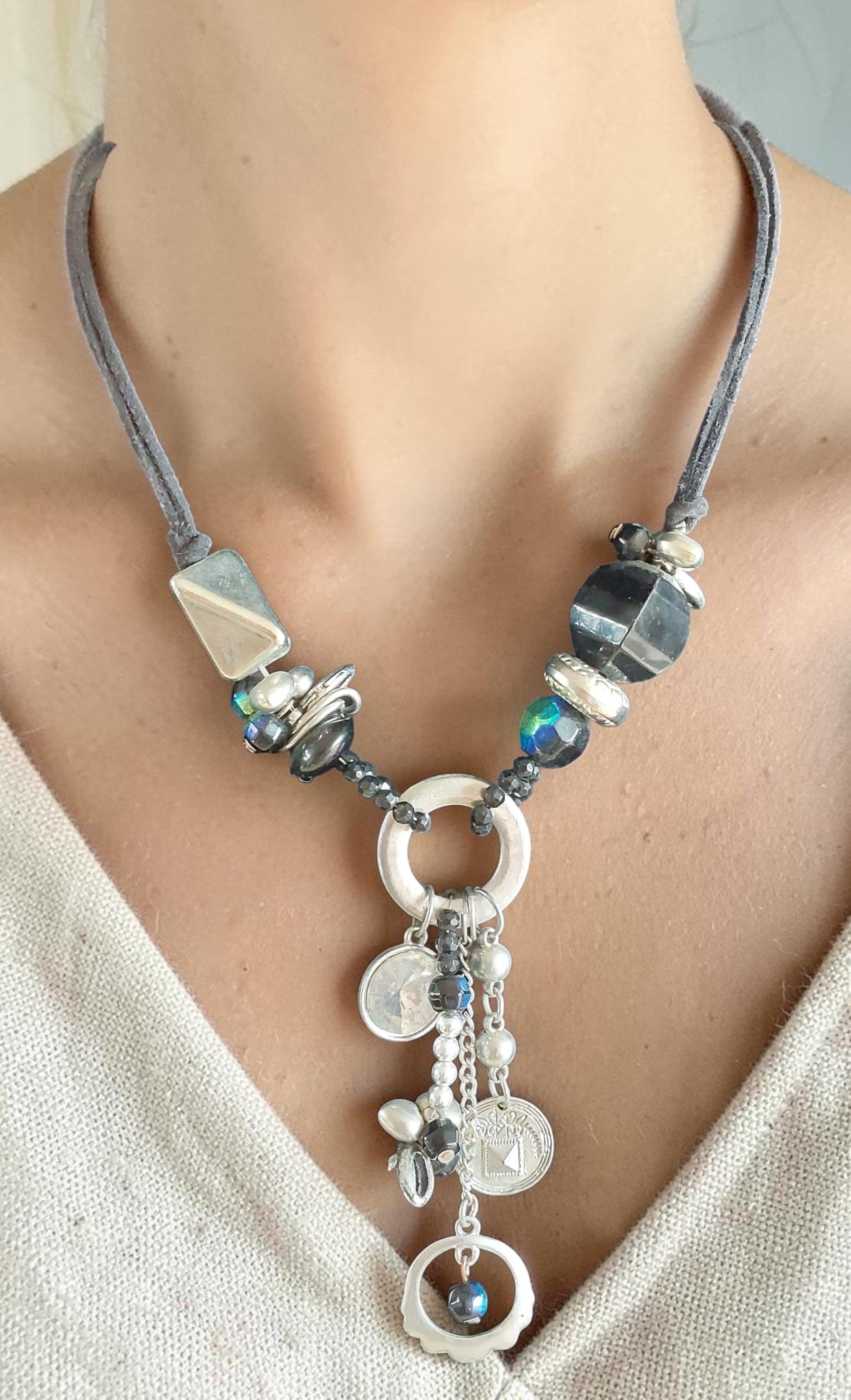 Halsketting met grijs suède koord, blauwe kristallen en facetgeslepen hematietkralen, afgewerkt met een cascade van zilveren ringen, muntjes en een blauwe steenhanger.