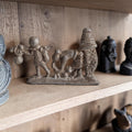 Grijs metalen tafereel met een figuur die goederen draagt, een voorovergebogen figuur en een kegelvormige hut of graanspieker.
