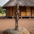 Handgesneden Afrikaans voorouderbeeld van donker hout met een gestileerd hoofd en een staande menselijke figuur op een cilindrische sokkel.
