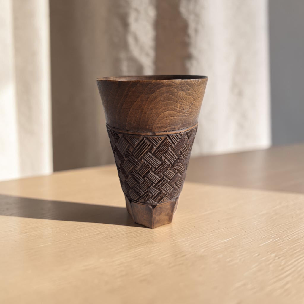 Handgesneden Kuba palmwijnbeker van warmbruin hardhout met geometrisch vlechtwerk in hoogreliëf, een gladde bovenrand en een achthoekige voet.
