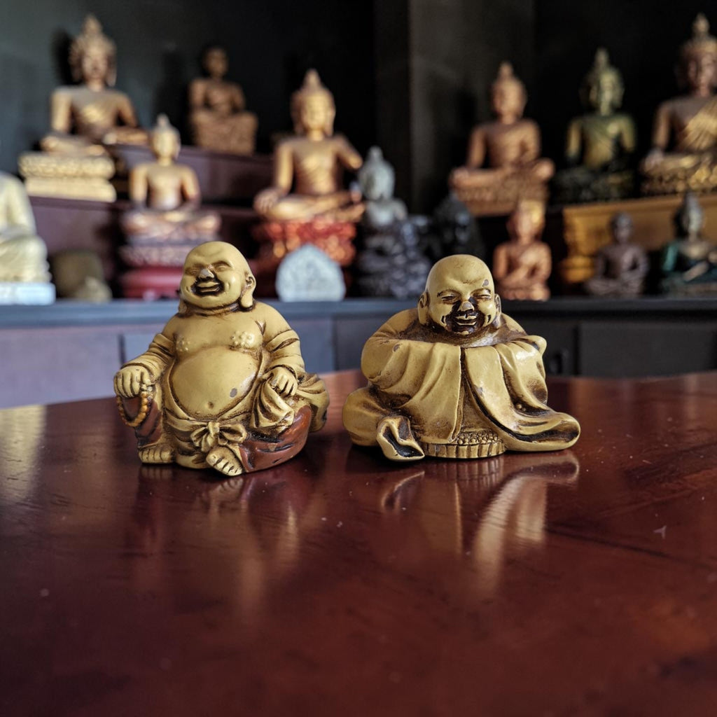 Set van twee zittende Lachende Boeddha beeldjes in goudgele kunsthars met donkere patina-accenten, hoogte ca. 3 cm.