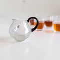 Leerdam glazen theepot met een bolle vorm, zwart handvat en gedeeltelijk aanwezige Leerdam-sticker.

