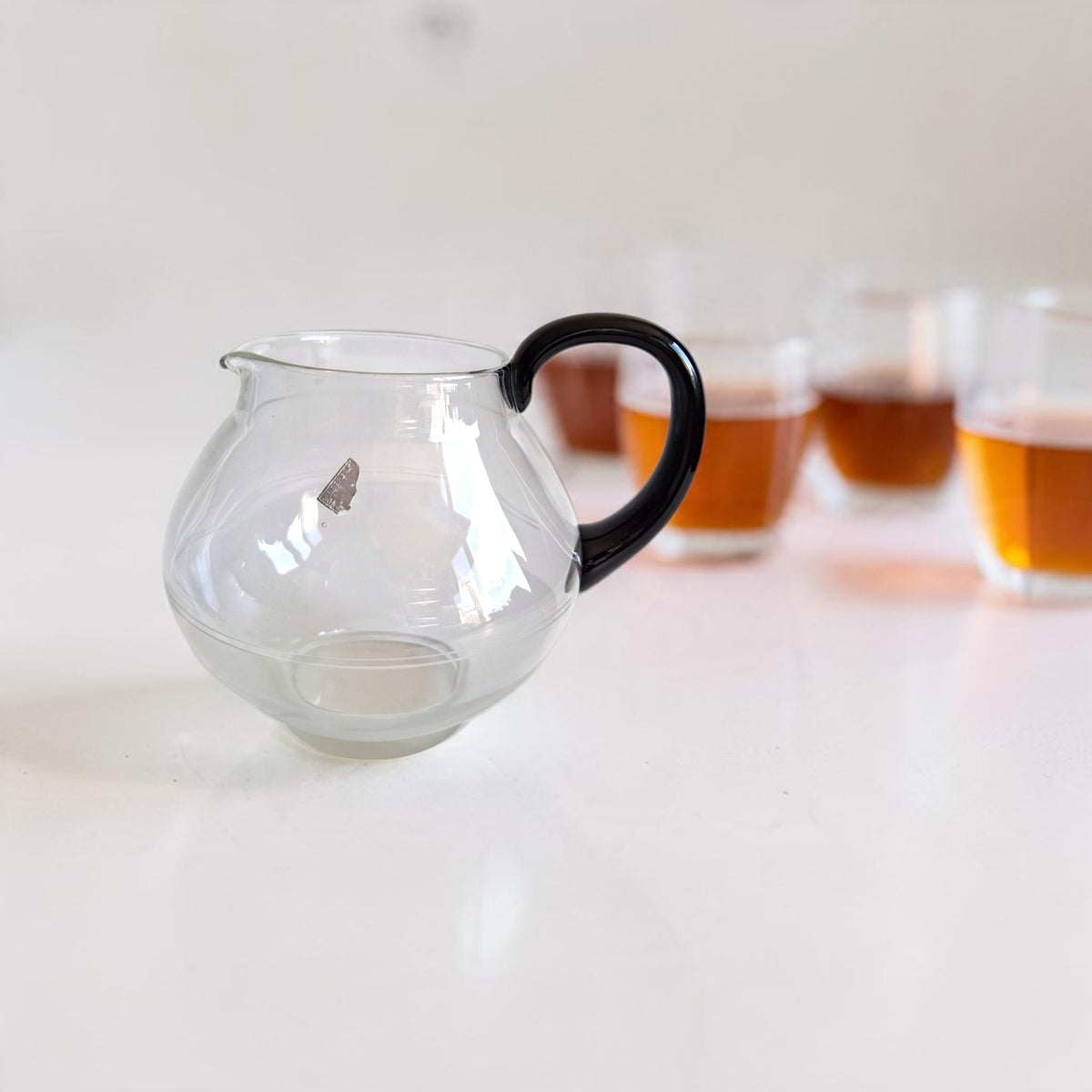 Leerdam glazen theepot met een bolle vorm, zwart handvat en gedeeltelijk aanwezige Leerdam-sticker.

