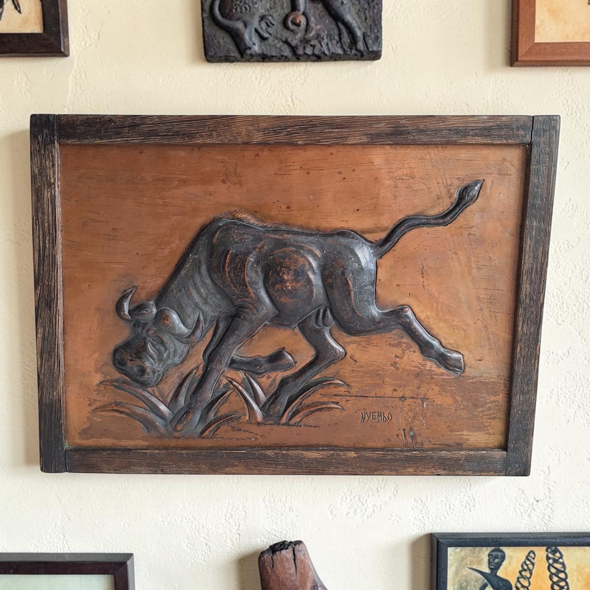 Ingelijst koperen reliëf van een Afrikaanse buffel die met gebogen kop door het gras stormt, rechtsonder gesigneerd NYEMBO en gedateerd 1980.
