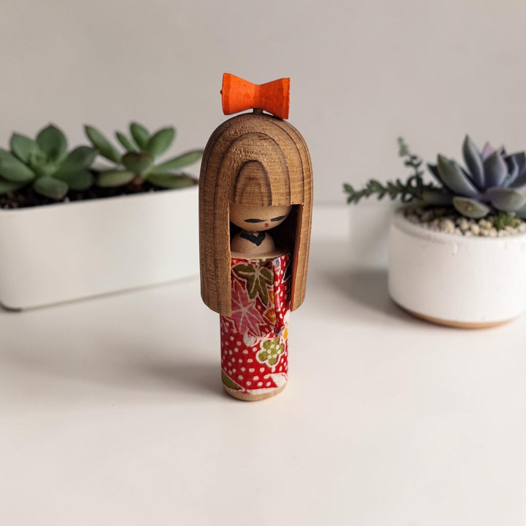 Miniatuur Sosaku Kokeshi met rood-wit cilindrisch lichaam en oranje strik op het hoofd, h8,5 x b3 x d3 cm, Japanse volkskunst.
